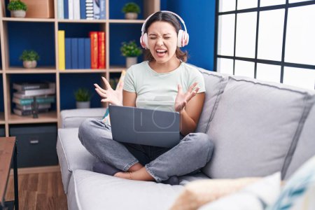 Foto de Hispanic young woman using laptop at home wearing headphones crazy and mad shouting and yelling with aggressive expression and arms raised. frustration concept. - Imagen libre de derechos