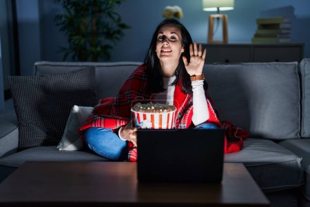 Téléchargez les photos : Hispanic woman eating popcorn watching a movie on the sofa waiving saying hello happy and smiling, friendly welcome gesture - en image libre de droit