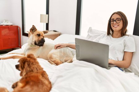 Téléchargez les photos : Jeune femme hispanique utilisant un ordinateur portable assis sur le lit avec des chiens dans la chambre - en image libre de droit