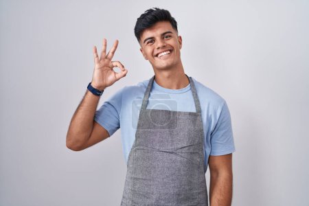 Foto de Joven hispano vistiendo delantal sobre fondo blanco sonriendo positivo haciendo signo de ok con la mano y los dedos. expresión exitosa. - Imagen libre de derechos