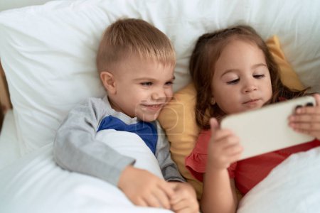 Foto de Adorable chica y niño viendo vídeo en el teléfono inteligente tumbado en la cama en el dormitorio - Imagen libre de derechos
