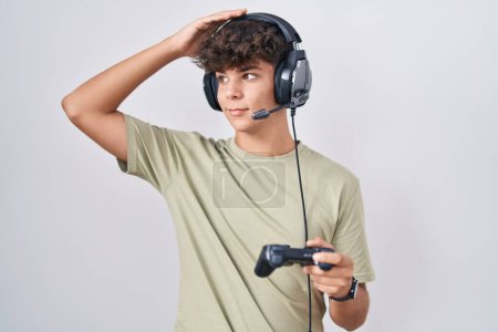 Téléchargez les photos : Adolescent hispanique jouant contrôleur de tenue de jeu vidéo souriant cheveux touchants confiants avec geste de la main vers le haut, posant attrayant et à la mode - en image libre de droit