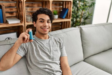 Téléchargez les photos : Jeune homme hispanique utilisant inhalateur assis sur le canapé à la maison. - en image libre de droit