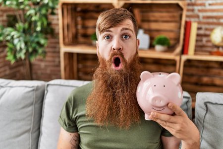 Photo for Redhead man holding piggy bank scared and amazed with open mouth for surprise, disbelief face - Royalty Free Image