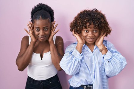 Téléchargez les photos : Two african women standing over pink background trying to hear both hands on ear gesture, curious for gossip. hearing problem, deaf - en image libre de droit