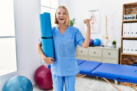 Photo for Young caucasian physiotherapist woman holding yoga mat pointing thumb up to the side smiling happy with open mouth - Royalty Free Image