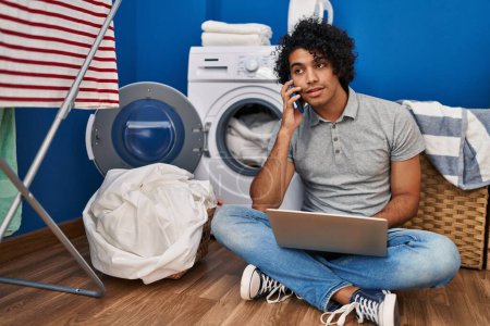 Téléchargez les photos : Jeune homme hispanique utilisant un ordinateur portable et parlant sur le smartphone attendant la machine à laver à la buanderie - en image libre de droit