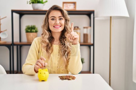 Photo for Young caucasian woman sitting on the table with piggy bank and coins smiling with an idea or question pointing finger with happy face, number one - Royalty Free Image
