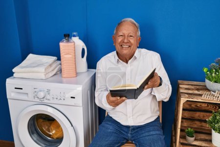 Téléchargez les photos : Senior man reading book waiting for washing machine at laundry room - en image libre de droit