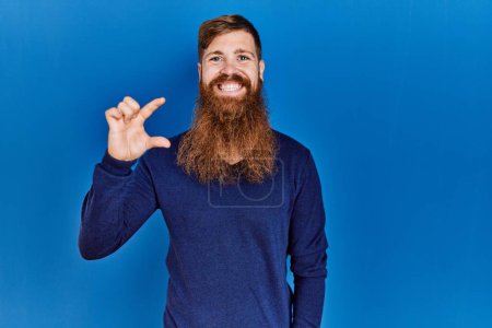 Téléchargez les photos : Homme rousse à longue barbe portant un pull bleu décontracté sur fond bleu geste souriant et confiant avec la main faisant signe de petite taille avec les doigts regardant et la caméra. concept de mesure. - en image libre de droit