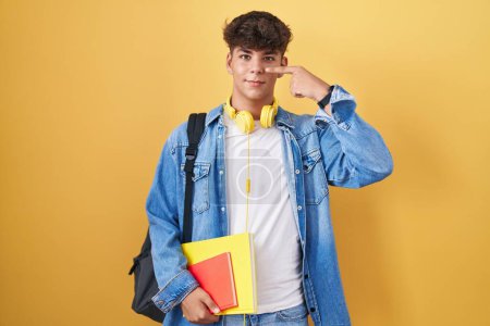 Foto de Adolescente hispana con mochila de estudiante y libros apuntando con el dedo de la mano a la cara y la nariz, sonriendo alegre. concepto de belleza - Imagen libre de derechos