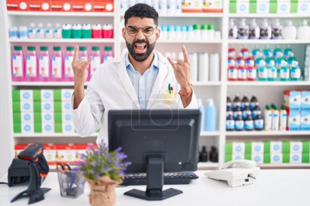 Téléchargez les photos : Hispanic man with beard working at pharmacy drugstore shouting with crazy expression doing rock symbol with hands up. music star. heavy concept. - en image libre de droit