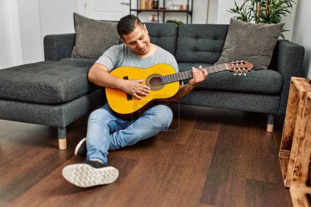 Foto de Joven hombre hispano sonriendo feliz tocando la guitarra clásica sentado en el suelo en casa. - Imagen libre de derechos
