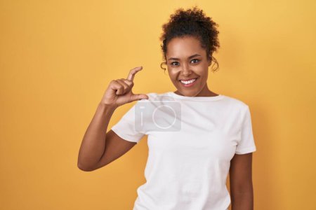 Photo for Young hispanic woman with curly hair standing over yellow background smiling and confident gesturing with hand doing small size sign with fingers looking and the camera. measure concept. - Royalty Free Image