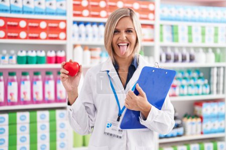 Téléchargez les photos : Jeune femme travaillant à la pharmacie drugstore tenant coeur langue collante heureux avec expression drôle. - en image libre de droit