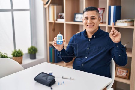 Téléchargez les photos : Jeune homme hispanique tenant dispositif glucomètre surpris par une idée ou une question pointant du doigt avec le visage heureux, numéro un - en image libre de droit