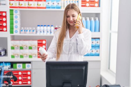 Foto de Joven mujer caucásica farmacéutica sosteniendo pastillas botella hablando en el teléfono inteligente en la farmacia - Imagen libre de derechos