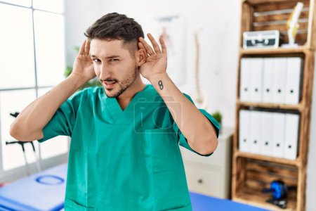 Foto de Joven fisioterapeuta hombre que trabaja en la clínica de recuperación del dolor tratando de escuchar ambas manos en el gesto del oído, curioso por chismes. problema auditivo, sordo - Imagen libre de derechos