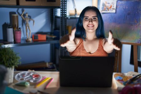 Photo for Young modern girl with blue hair sitting at art studio with laptop at night approving doing positive gesture with hand, thumbs up smiling and happy for success. winner gesture. - Royalty Free Image