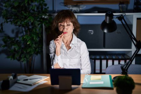 Téléchargez les photos : Femme d'âge moyen travaillant au bureau la nuit bouche et lèvres fermées comme zip avec les doigts. secret et silencieux, tabou parlant - en image libre de droit