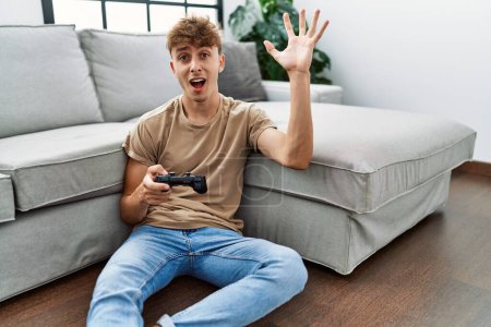 Photo for Young caucasian man playing video game holding controller at home celebrating victory with happy smile and winner expression with raised hands - Royalty Free Image