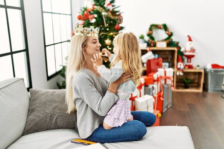 Foto de Mother and daughter wearing queen cronw hugging each other sitting by christmas tree at home - Imagen libre de derechos