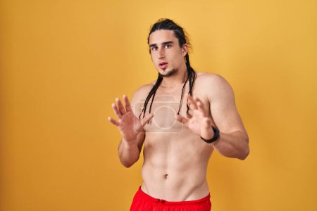 Foto de Hispanic man with long hair standing shirtless over yellow background afraid and terrified with fear expression stop gesture with hands, shouting in shock. panic concept. - Imagen libre de derechos
