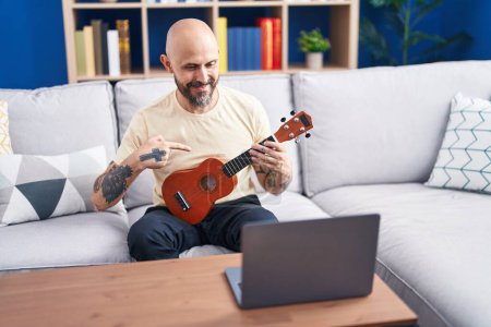 Téléchargez les photos : Jeune homme chauve ayant en ligne ukulele classe assis sur le canapé à la maison - en image libre de droit