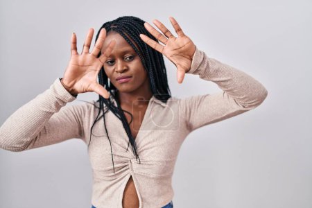 Foto de Mujer africana con trenzas de pie sobre fondo blanco haciendo marco usando las manos palmas y los dedos, perspectiva de la cámara - Imagen libre de derechos