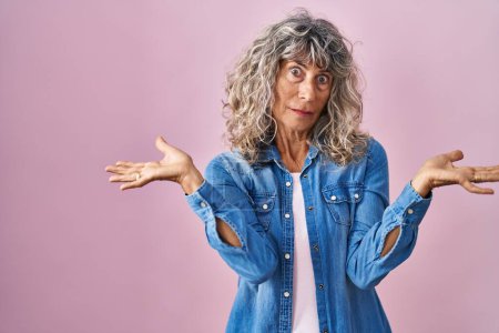 Photo for Middle age woman standing over pink background clueless and confused expression with arms and hands raised. doubt concept. - Royalty Free Image