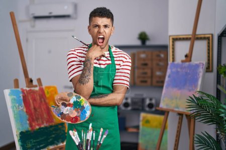 Foto de Joven brasileño sosteniendo paleta de pintores en el estudio del artista enojado y loco gritando frustrado y furioso, gritando con ira. rabia y concepto agresivo. - Imagen libre de derechos