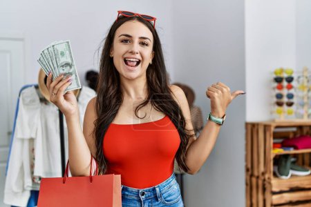 Foto de Joven morena sosteniendo bolsas de compras y dólares apuntando el pulgar hacia el lado sonriendo feliz con la boca abierta - Imagen libre de derechos