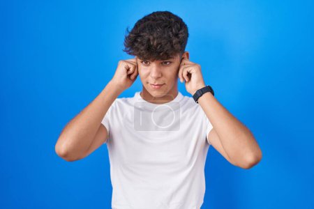 Téléchargez les photos : Adolescent hispanique debout sur fond bleu couvrant les oreilles avec des doigts avec une expression agacée pour le bruit de la musique forte. concept sourd. - en image libre de droit
