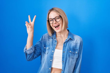 Foto de Young caucasian woman standing over blue background smiling with happy face winking at the camera doing victory sign. number two. - Imagen libre de derechos
