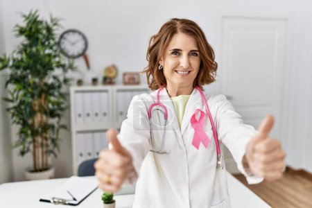 Foto de Médico de mediana edad mujer con cinta de cáncer rosa en uniforme que aprueba hacer gesto positivo con la mano, pulgares hacia arriba sonriendo y feliz por el éxito. gesto ganador. - Imagen libre de derechos