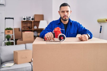Photo for Hispanic man working on moving holding packing tape thinking attitude and sober expression looking self confident - Royalty Free Image