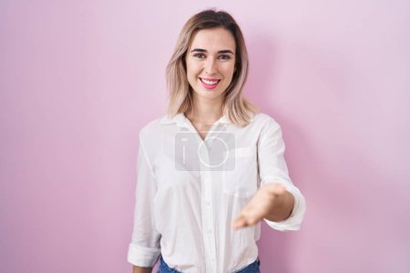 Téléchargez les photos : Young beautiful woman standing over pink background smiling cheerful offering palm hand giving assistance and acceptance. - en image libre de droit