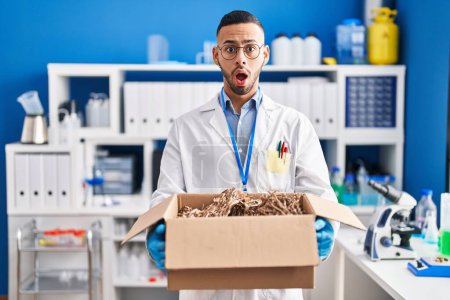 Photo for Young hispanic man working at scientist laboratory holding cardboard box afraid and shocked with surprise and amazed expression, fear and excited face. - Royalty Free Image