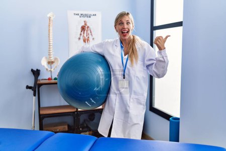 Photo for Beautiful woman holding pilates ball at physiotherapy clinic pointing thumb up to the side smiling happy with open mouth - Royalty Free Image