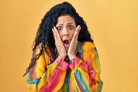 Photo for Middle age hispanic woman standing over yellow background afraid and shocked, surprise and amazed expression with hands on face - Royalty Free Image
