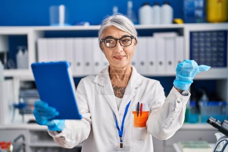 Foto de Mujer de mediana edad con el pelo gris trabajando en el laboratorio científico haciendo videollamada apuntando el pulgar hacia el lado sonriendo feliz con la boca abierta - Imagen libre de derechos