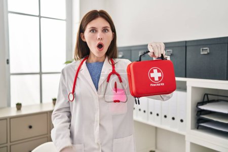 Foto de Joven doctora sosteniendo botiquín de primeros auxilios asustada y sorprendida con la boca abierta para sorpresa, cara de incredulidad - Imagen libre de derechos
