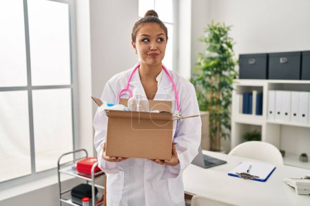 Photo for Young hispanic doctor holding box with medical items smiling looking to the side and staring away thinking. - Royalty Free Image