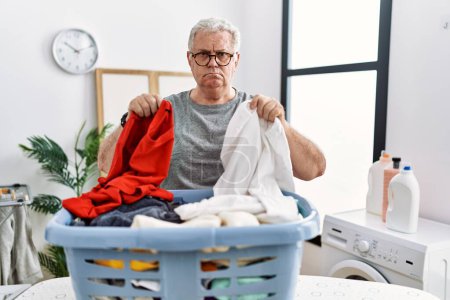 Photo for Senior caucasian man doing laundry holding clothes from wicker basket relaxed with serious expression on face. simple and natural looking at the camera. - Royalty Free Image