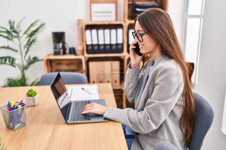 Photo for Young beautiful hispanic woman business worker using laptop talking on smartphone at office - Royalty Free Image