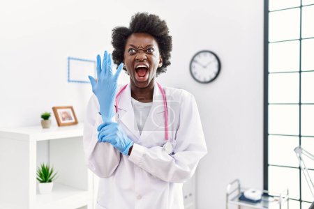 Foto de Médico africano mujer usando guantes de látex en la clínica médica enojado y loco gritando frustrado y furioso, gritando con ira. rabia y concepto agresivo. - Imagen libre de derechos