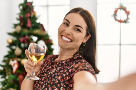 Téléchargez les photos : Jeune belle femme hispanique faire selfie par caméra debout près de l'arbre de Noël à la maison - en image libre de droit