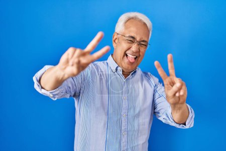 Foto de Hombre mayor hispano con gafas sonriendo con la lengua hacia fuera mostrando los dedos de ambas manos haciendo signo de victoria. número dos. - Imagen libre de derechos