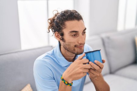 Téléchargez les photos : Jeune homme hispanique buvant du café assis sur le canapé à la maison - en image libre de droit