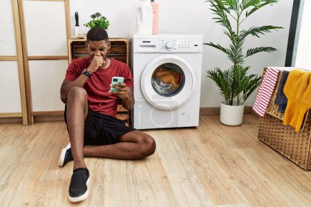 Foto de Hombre afroamericano joven usando un teléfono inteligente esperando que la lavadora se sienta mal y tose como síntoma de resfriado o bronquitis. concepto de atención sanitaria. - Imagen libre de derechos